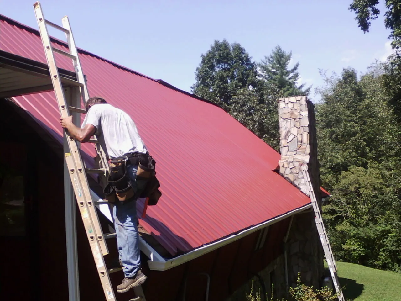 Roofer on a ladder beside a red metal roof and stone chimney — Soffit & Fascia Repair by Roofing Brothers in Harker Heights